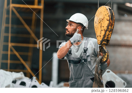 With crane hook. Young factory worker in grey uniform With crane hook. Young factory worker in grey uniform 128596862