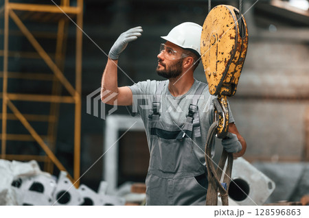 With crane hook. Young factory worker in grey uniform With crane hook. Young factory worker in grey uniform 128596863