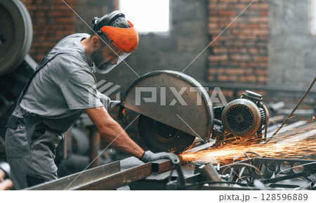 Side view, cutting the metal. Young factory worker in grey uniform 128596889