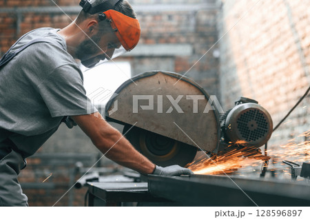 Focused at work, cutting the metal. Young man at factory in grey uniform Focused at work, cutting the metal. Young man at factory in grey uniform 128596897