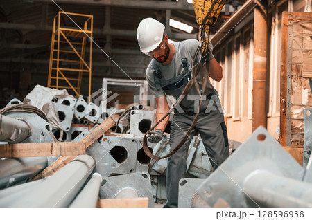 Standing and using crane hook. Young factory worker in grey uniform 128596938
