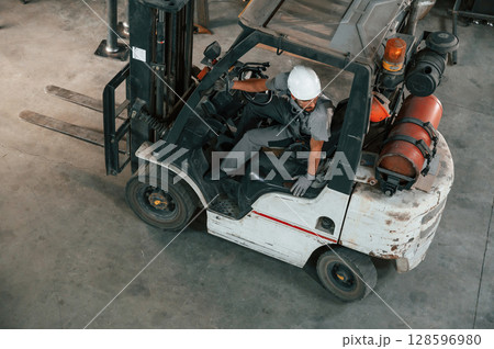 View from the top, in the forklift. Young factory worker in grey uniform View from the top, in the forklift. Young factory worker in grey uniform 128596980