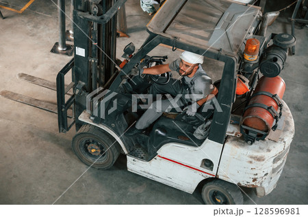 View from the top, in the forklift. Young factory worker in grey uniform View from the top, in the forklift. Young factory worker in grey uniform 128596981