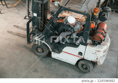 View from the top, in the forklift. Young factory worker in grey uniform View from the top, in the forklift. Young factory worker in grey uniform 128596982