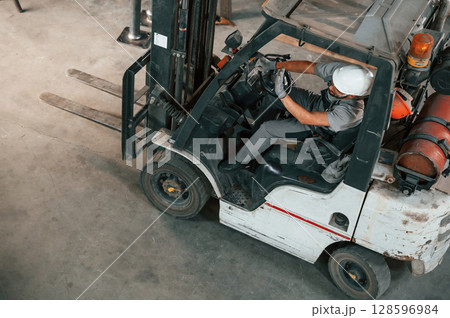 View from the top, in the forklift. Young factory worker in grey uniform View from the top, in the forklift. Young factory worker in grey uniform 128596984