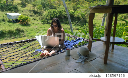 A young woman enjoys her freelance work from a hammock, nestled on a tropical terrace, surrounded by lush rice fields and vibrant jungle  128597126