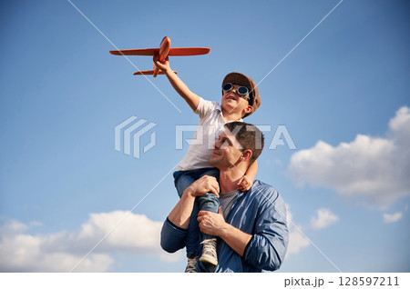 Playing with toy plane, sitting on the shoulders. Father and little son are having fun outdoors Playing with toy plane, sitting on the shoulders. Father and little son are having fun outdoors 128597211