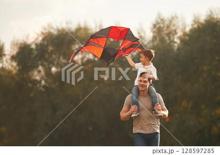 Red and black colored kite in hands. Father and little son are playing and having fun outdoors Red and black colored kite in hands. Father and little son are playing and having fun outdoors 128597285