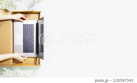 Buying and delivering new laptop. Romantic overhead shot of womans hands holding laptop in cardboard box on blue background with white spring flowers. Flat lay, copy space Buying and delivering new laptop. Romantic overhead shot of womans hands holding laptop in cardboard box on blue background with white spring flowers. Flat lay, copy space 128597342