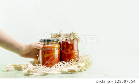 Probiotic foods. Sustainable still life with woman hand hold pickled or fermented vegetables. Lecho and ajvar in glass jars in mesh bag on blue background. Home food preserving or canning. Copy space Probiotic foods. Sustainable still life with woman hand hold pickled or fermented vegetables. Lecho and ajvar in glass jars in mesh bag on blue background. Home food preserving or canning. Copy space 128597354