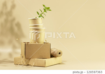 Sustainable still life. Eco friendly packaging concept. Cardboard boxes, disposable cups with green shoot on beige background. Concept of ecology, conservation, plastic free and recycling. Copy space 128597363