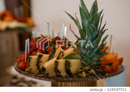 Vibrant Fruit Platter Display with Pineapple, Strawberries, Oranges, and Skewers on Elegant Glass Stand, Bright Tropical Color Palette. Vibrant Fruit Platter Display with Pineapple, Strawberries, Oranges, and Skewers on Elegant Glass Stand, Bright Tropical Color Palette. 128597551