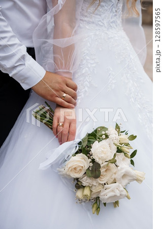 Romantic Wedding Rings Exchange: Close-up of Bride and Grooms Hands with Gold Bands, White Bouquet, Elegant Dress and Classic Marriage. 128597565