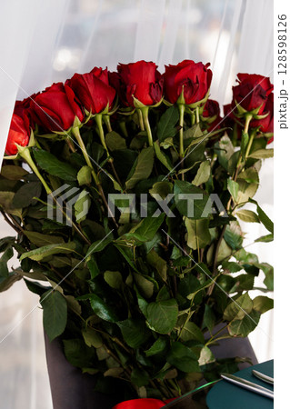 Vibrant Red Roses Bouquet in Brown Vase. Close-Up of Long-Stemmed Flowers. Romantic Gesture. Lush Floral Arrangement. Dark Green Foliage. 128598126