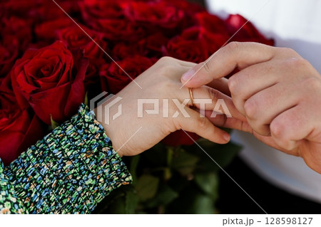 Romantic Engagement with Red Roses: Close-Up of Hands Exchanging Rings Amidst Vibrant Flower Bouquet, Love, Commitment, and Celebration. 128598127