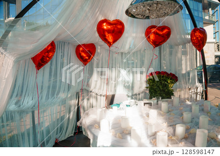 Romantic Valentine's Day Indoor Setup with Red Heart Balloons, White Drapery, Roses, Candles, and a Cozy Table for a Couple. 128598147