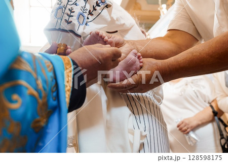 Orthodox Christian Baptism Ceremony with Priest Anointing Infant's Feet, White Robes, Spiritual Ritual, Blue and Gold Vestments. Orthodox Christian Baptism Ceremony with Priest Anointing Infant's Feet, White Robes, Spiritual Ritual, Blue and Gold Vestments. 128598175