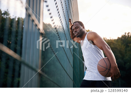 Leaning on the metal construction. Young black man is with basketball ball outdoors Leaning on the metal construction. Young black man is with basketball ball outdoors 128599140