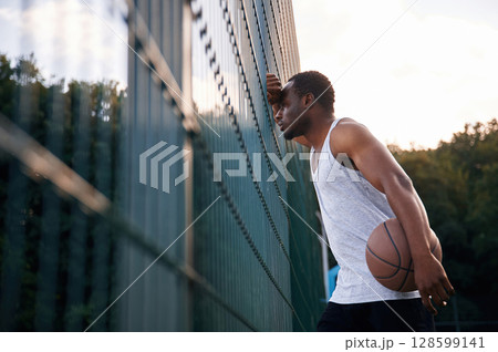 Leaning on the metal construction. Young black man is with basketball ball outdoors Leaning on the metal construction. Young black man is with basketball ball outdoors 128599141
