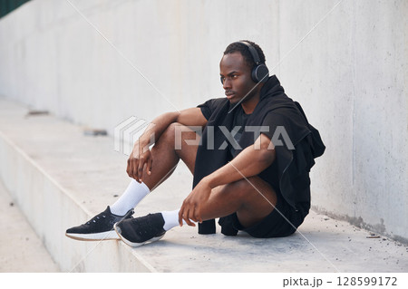 Sitting by the white wall. Handsome black man is posing outdoors at daytime Sitting by the white wall. Handsome black man is posing outdoors at daytime 128599172