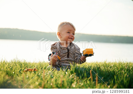 Against lake behind. Cute little boy is having fun outdoors on the ground with green grass 128599181