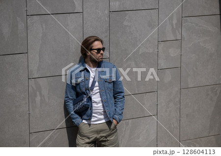 A stylish young man in a denim jacket, white T-shirt, and shorts poses confidently against a gray wall A stylish young man in a denim jacket, white T-shirt, and shorts poses confidently against a gray wall 128604113