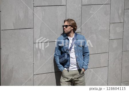 A stylish young man in a denim jacket, white T-shirt, and shorts poses confidently against a gray wall 128604115