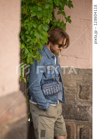 A stylish young man in a denim jacket and sunglasses poses confidently against a rustic wall A stylish young man in a denim jacket and sunglasses poses confidently against a rustic wall 128604118