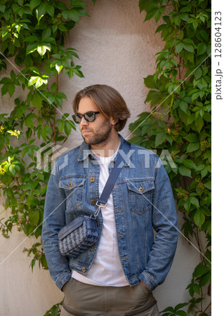 A stylish young man in a denim jacket and sunglasses poses confidently against a rustic wall 128604123