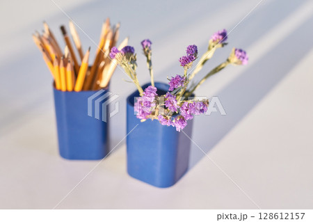 Purple flowers and pencils in blue containers on white surface with soft shadows Purple flowers and pencils in blue containers on white surface with soft shadows 128612157
