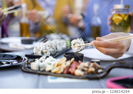 Close-up of a hand using chopsticks to eat sushi from a table set with other food and drinks. Friends enjoy the meal. 128614284