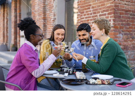 A diverse group of friends enjoys sushi outdoors, sharing food and laughter around a table. 128614291