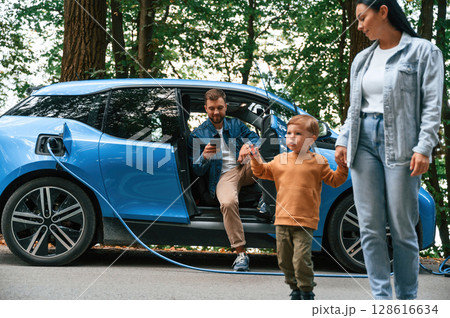 Walking forward. Mother, father and little son are waiting for electric car to charge outdoors 128616634