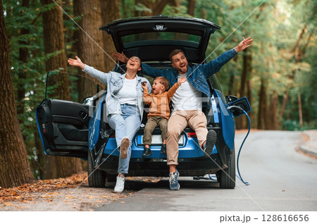 Sitting on the back of a vehicle. Mother, father and little son are waiting for electric car to charge outdoors 128616656