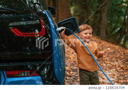 Holding the wire. Cute young boy is charging the blue electric car outdoors 128616704