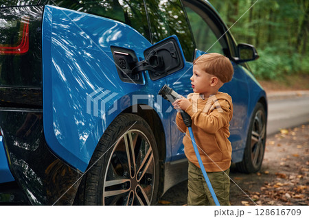 On the road. Cute young boy is charging the blue electric car outdoors 128616709