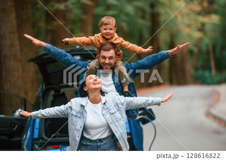 Playing together. Mother, father and little son are waiting for electric car to charge outdoors Playing together. Mother, father and little son are waiting for electric car to charge outdoors 128616822