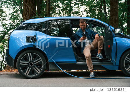 Sitting with doors opened. Handsome man is waiting for his electric car to charge, outdoors 128616854