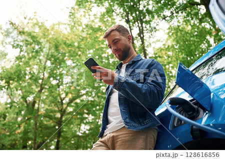 View from below, with smartphone. Handsome man is waiting for his electric car to charge, outdoors 128616856