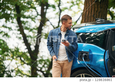 Standing and holding smartphone. Handsome man is waiting for his electric car to charge, outdoors Standing and holding smartphone. Handsome man is waiting for his electric car to charge, outdoors 128616861