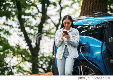 Woman with smartphone is standing near electric car that is charging outdoors Woman with smartphone is standing near electric car that is charging outdoors 128616900