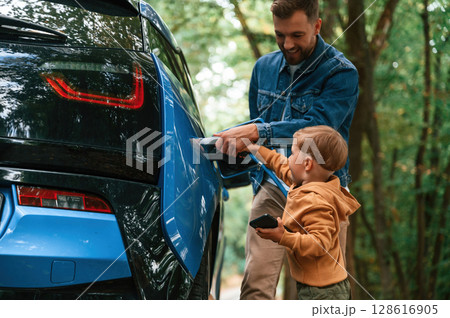 Teaching the boy where to put the wire. Father with his little son are waiting for electric car to charge Teaching the boy where to put the wire. Father with his little son are waiting for electric car to charge 128616905