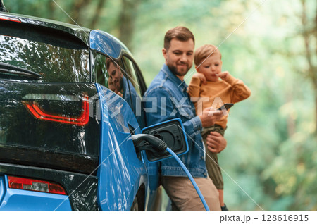 Holding boy in hands. Father with his little son are waiting for electric car to charge Holding boy in hands. Father with his little son are waiting for electric car to charge 128616915