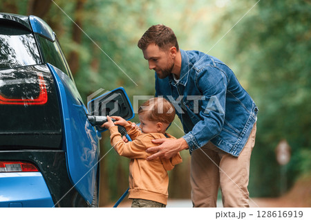 Teaching the boy where to put the wire. Father with his little son are waiting for electric car to charge Teaching the boy where to put the wire. Father with his little son are waiting for electric car to charge 128616919