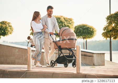 Little green trees behind. A young couple with a baby pram is walking together 128617113