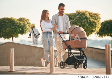Little green trees behind. A young couple with a baby pram is walking together Little green trees behind. A young couple with a baby pram is walking together 128617115