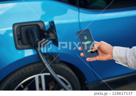 Charging process on the display of smartphone. Man with blue electric car on the station Charging process on the display of smartphone. Man with blue electric car on the station 128617137