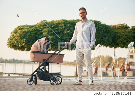 Young father is outdoors, standing with pram Young father is outdoors, standing with pram 128617141