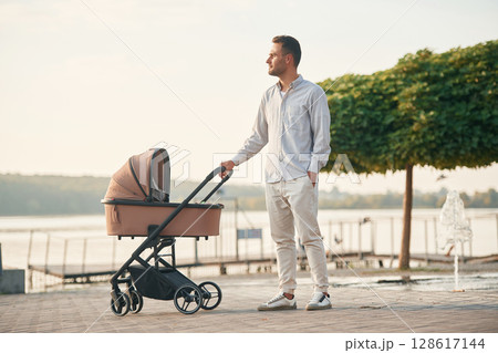 Green tree behind. Young father is outdoors, standing with pram Green tree behind. Young father is outdoors, standing with pram 128617144