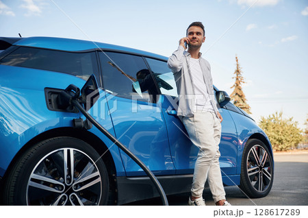 Talking by phone. Man with blue electric car on the charge station Talking by phone. Man with blue electric car on the charge station 128617289